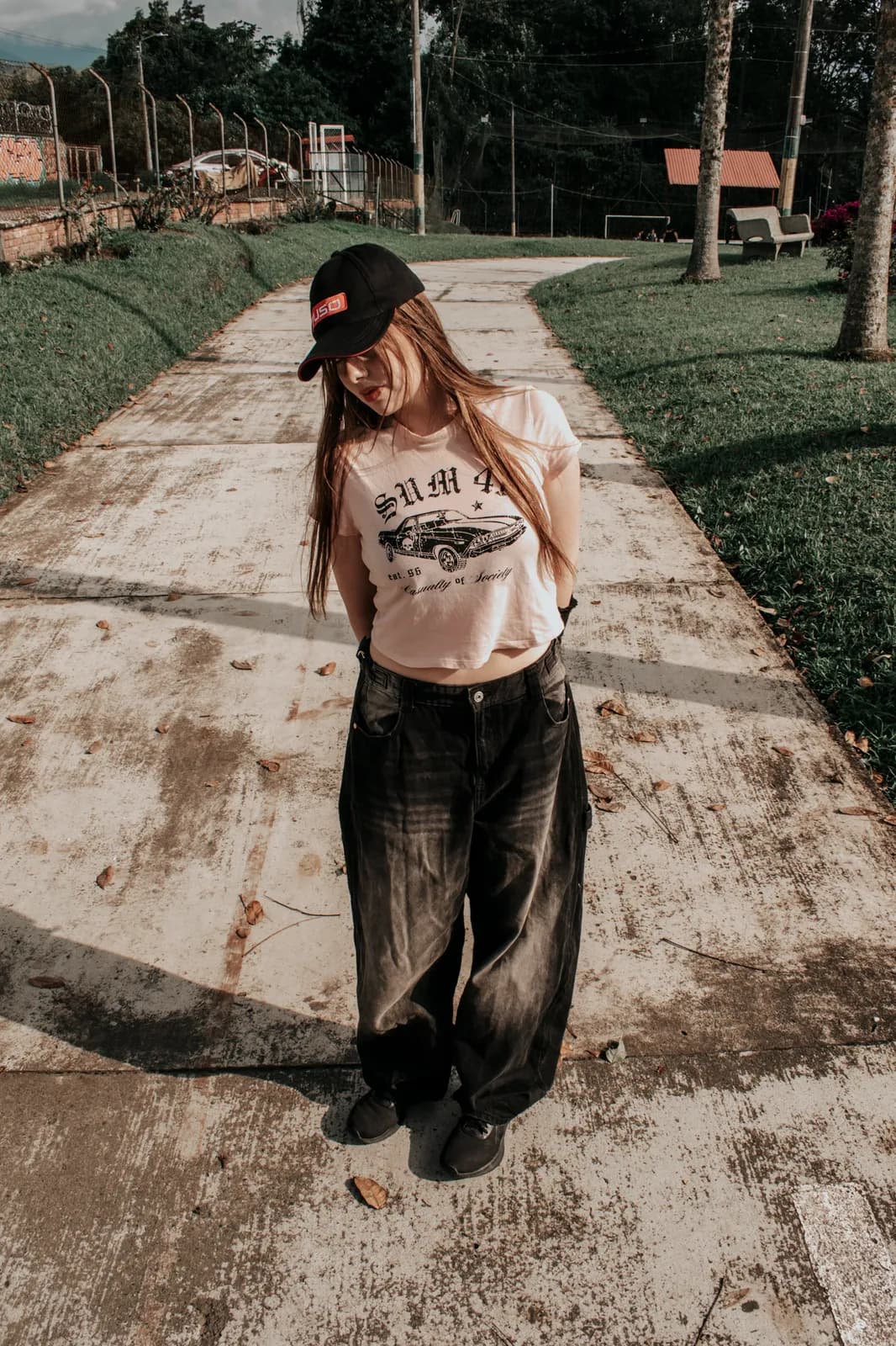 Joven con gorra y jeans anchos posando en un sendero arbolado