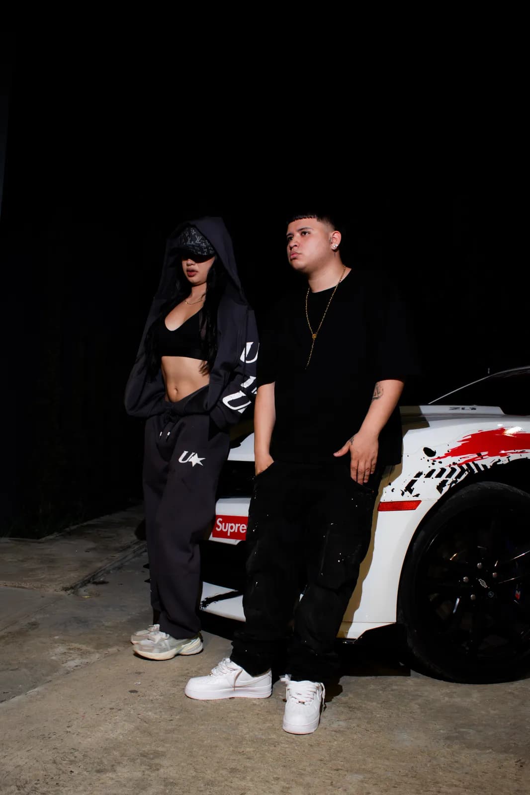 Dos jóvenes con ropa oscura posando frente a un carro deportivo de noche